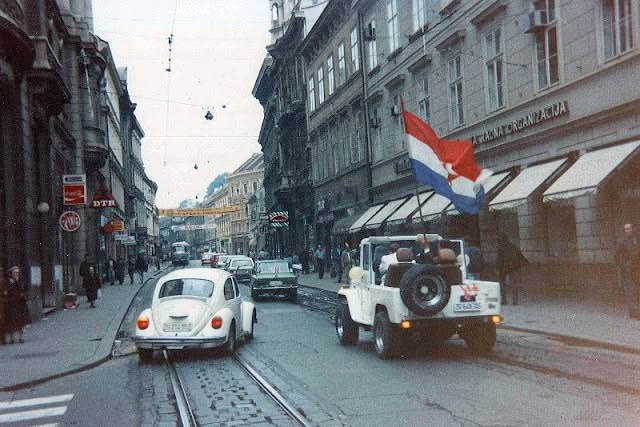 A street with tram tracks, parked cars and a vehicle flying a flag
